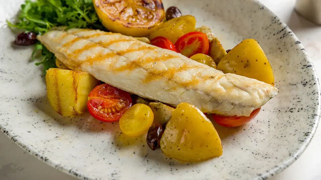 Filetto di persico grigliato su un piatto con patate arrosto, pomodorini, olive e limone caramellato.