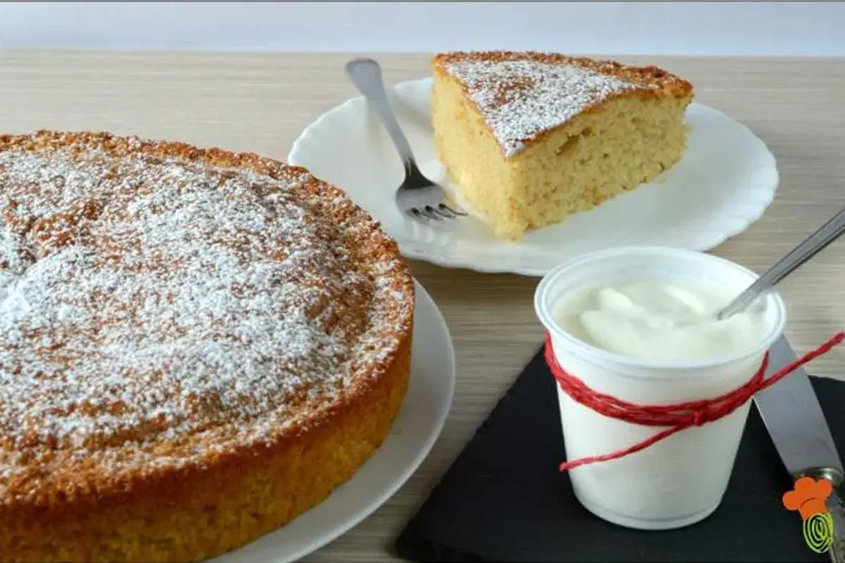 Torta 7 vasetti soffice e profumata, spolverata di zucchero a velo. Una fetta invitante e una coppetta di yogurt completano la scena.