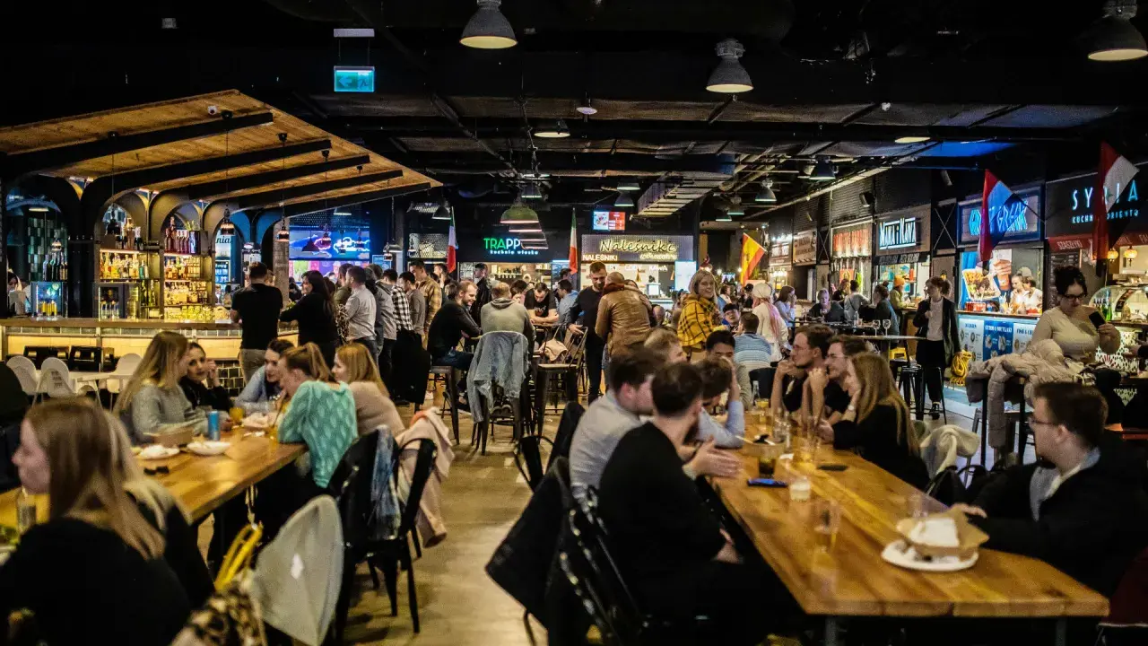 Tętniący życiem food hall Metropolia, gdzie ludzie jedzą i piją przy długich stołach, a w tle widać bary i stoiska z jedzeniem.