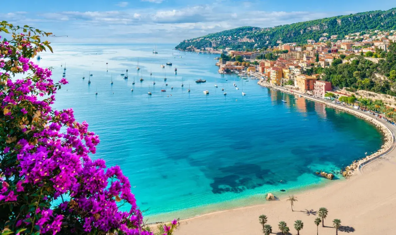 Blick auf einen malerischen Strand in Frankreich mit türkisfarbenem Wasser, Segelbooten und einer Küstenstadt, umrahmt von leuchtend lila Bougainvillea.