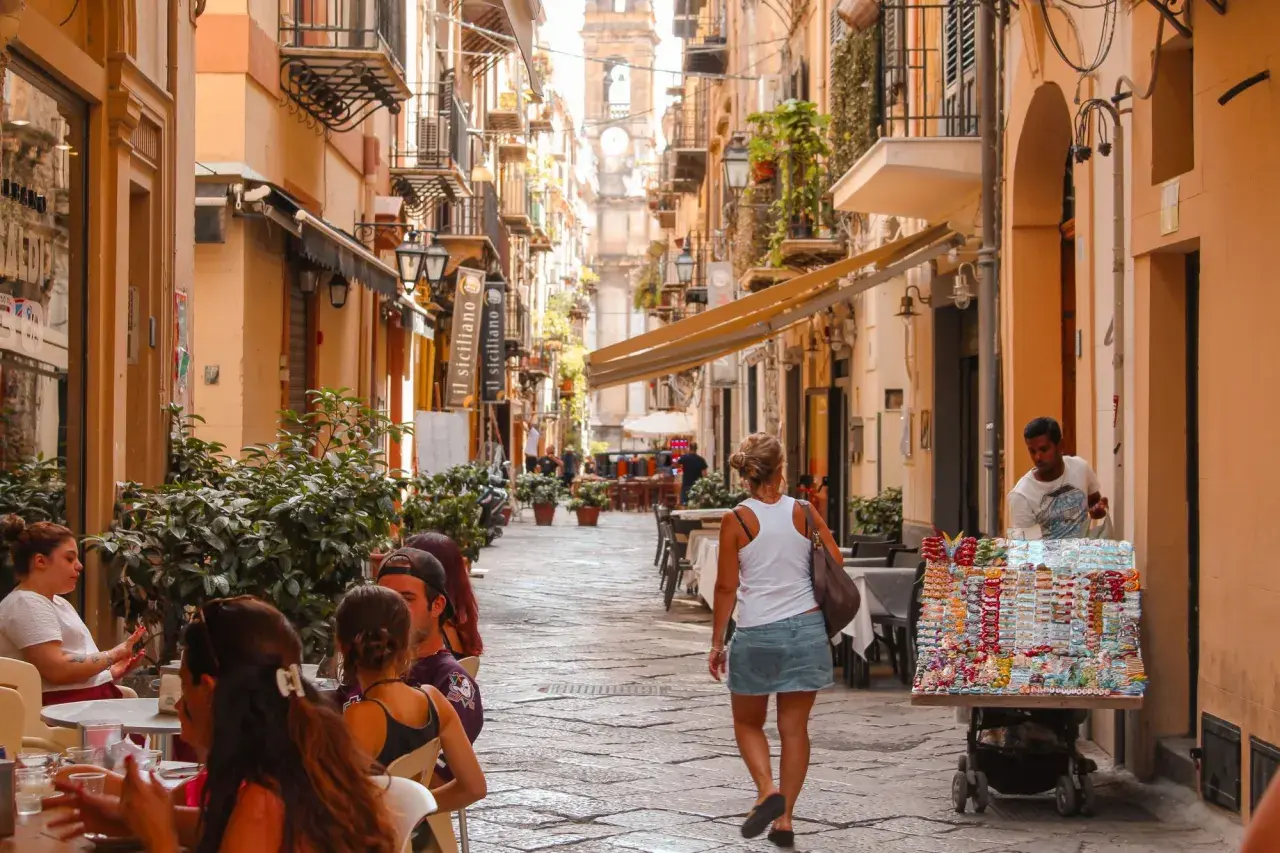 A woman walks down a bustling street in Sicily, past shops and cafes. The scene suggests a vibrant, lively atmosphere, answering the question: is Sicily safe for tourists to enjoy?