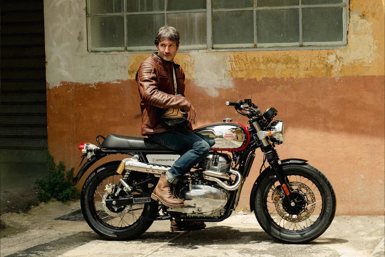 Uomo con giacca in pelle marrone seduto su una moto Royal Enfield Interceptor. I terminali moto cromati brillano sotto il sole.