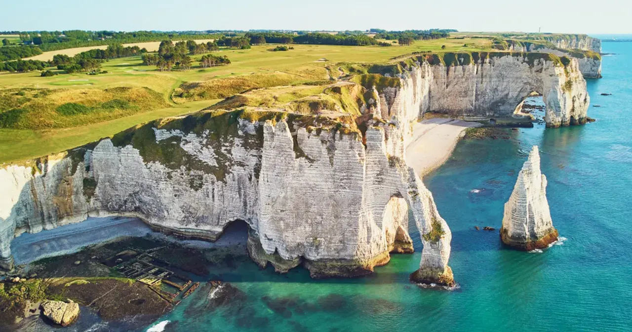 Wapienne klify nad kanałem La Manche, z łukiem skalnym i iglicą. Na szczycie klifów zielone pola golfowe.