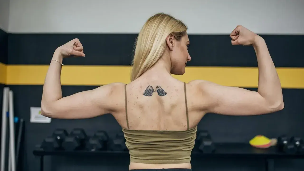 Silna kobieta z tatuażem skrzydeł na plecach napina biceps. Zastanawia się, ile po tatuażu nie można ćwiczyć, by go nie uszkodzić.