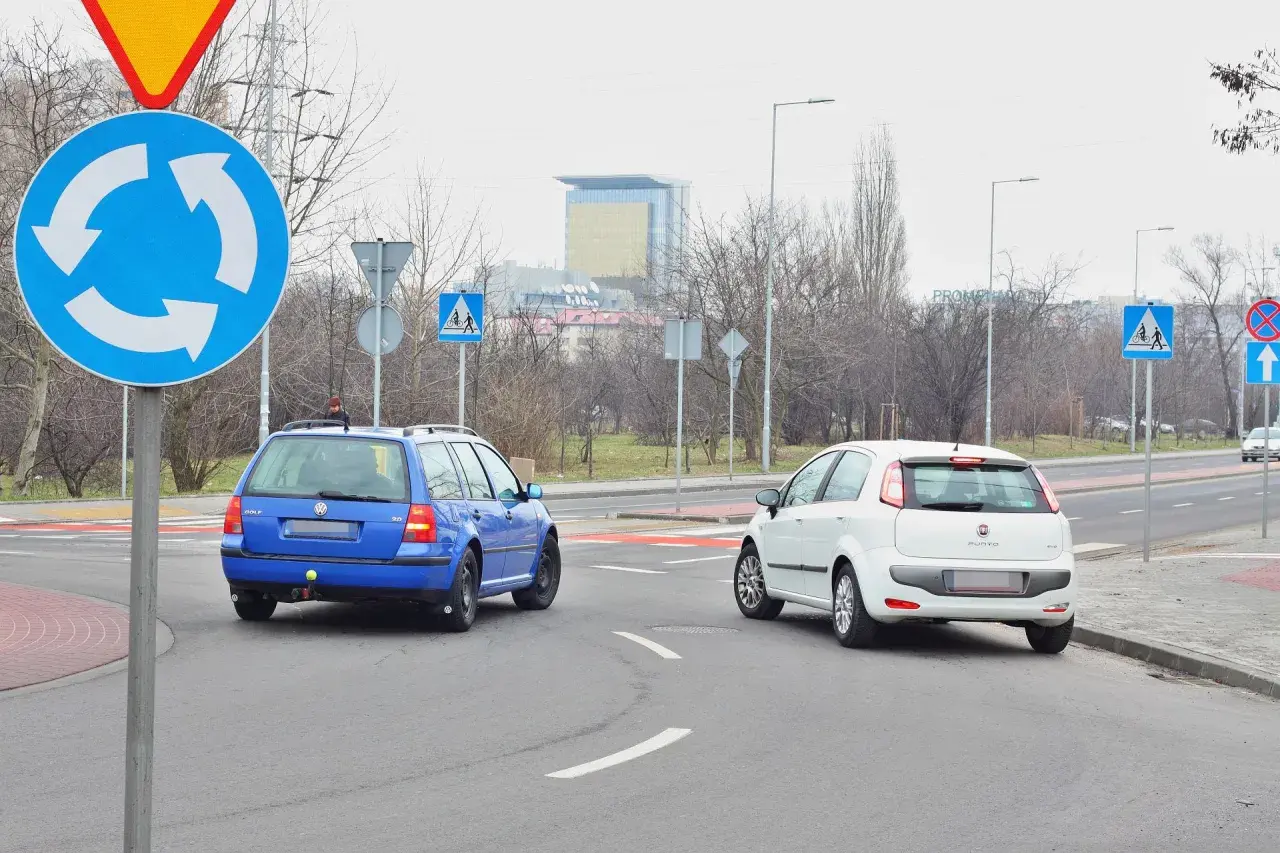 Niebieski Volkswagen i biały Fiat na rondzie. Kierowcy zastanawiają się, który zjazd z ronda na który pas wybrać, by bezpiecznie kontynuować jazdę.