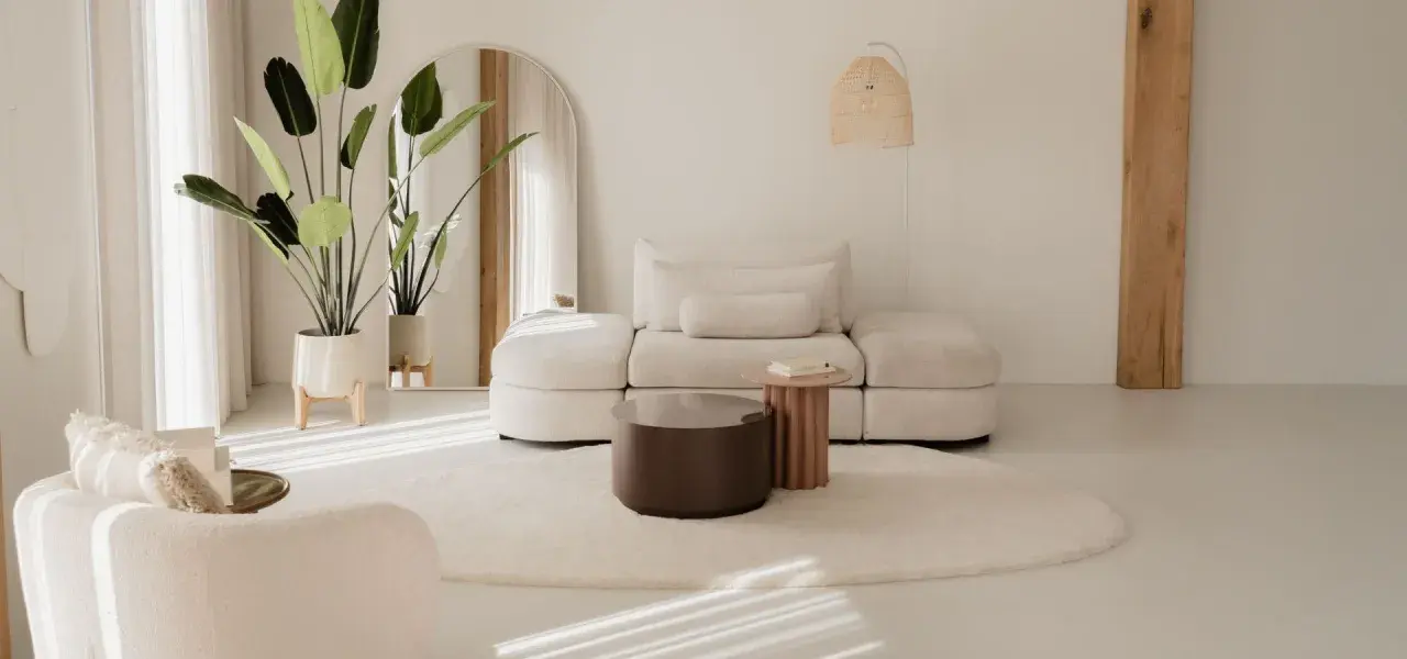 Ein helles Wohnzimmer im Japandi-Stil mit einem beigen Sofa, einem runden Teppich und einer großen Topfpflanze.