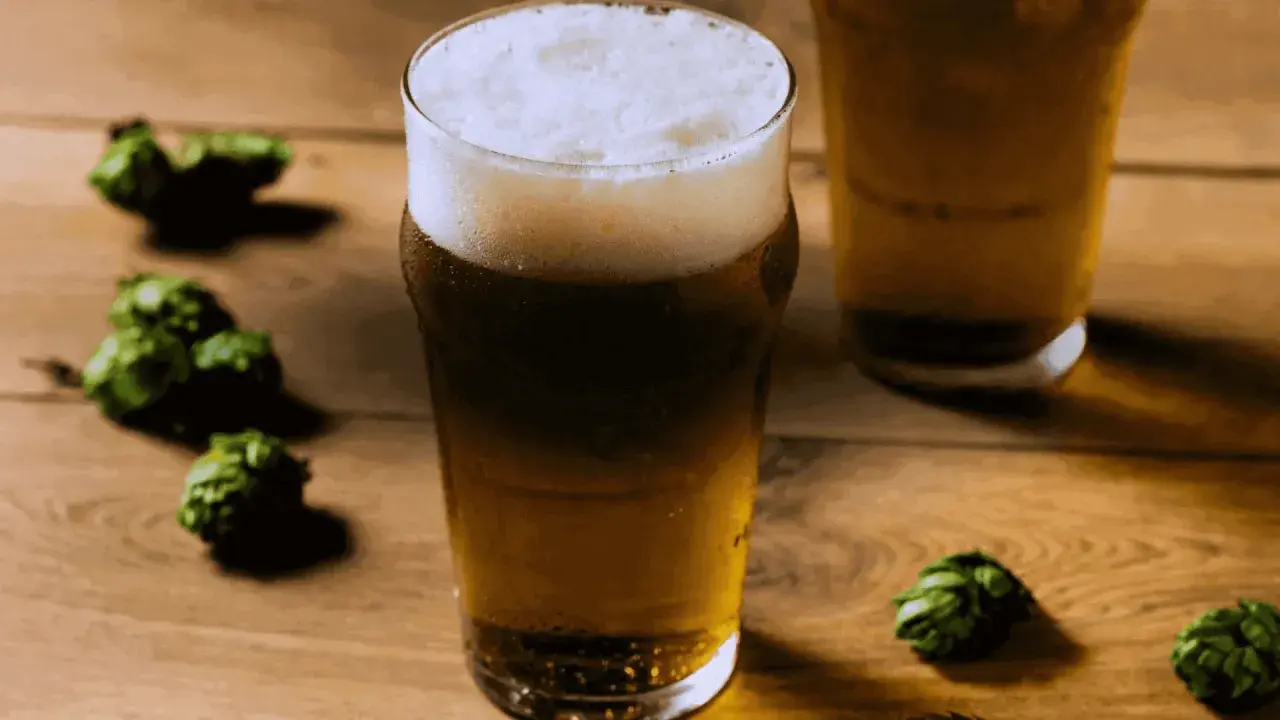 Bicchiere di birra dorata con schiuma bianca, circondato da luppoli verdi su un tavolo di legno. Perfetta per una dual IPA.