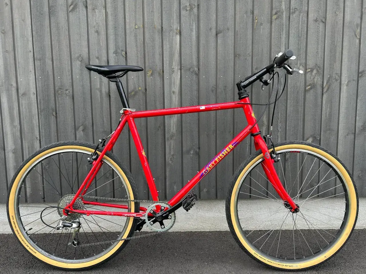 A vintage red Gary Fisher Advance mountain bike with tan tires stands against a wooden fence.