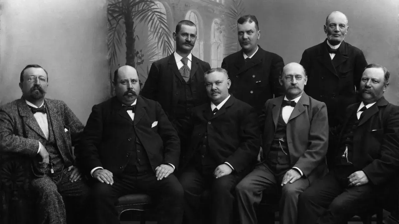 Gruppbild på män i 1900 talet kläder. De bär kostymer och flugor, redo för ett formellt porträtt.