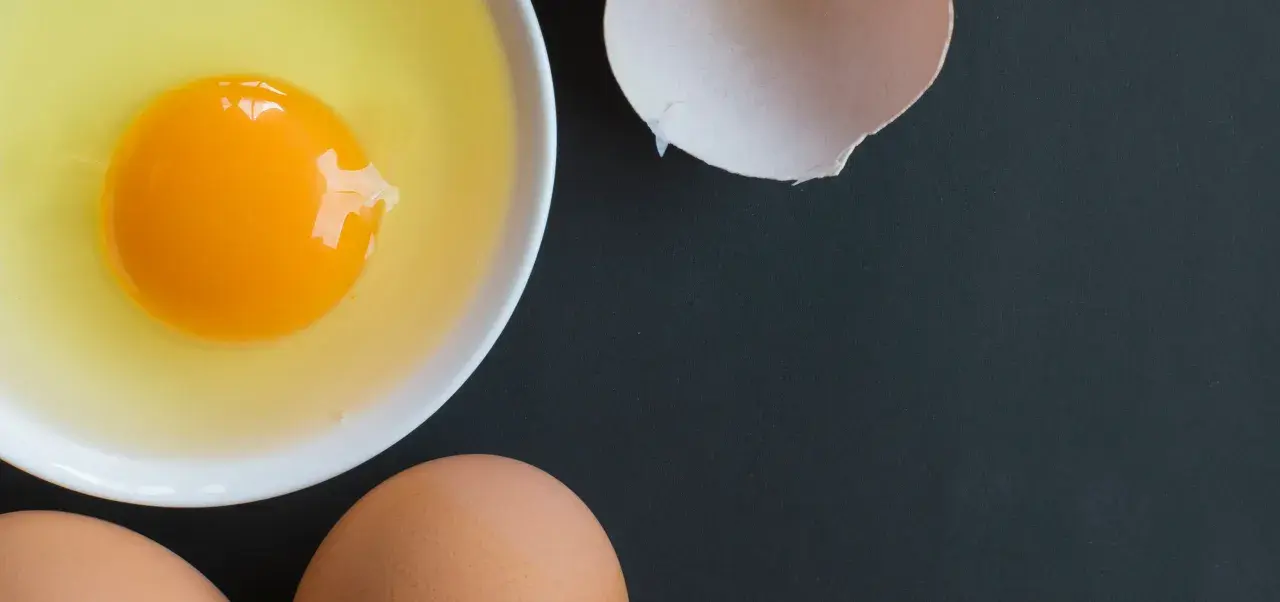 Un huevo crudo en un cuenco blanco, cáscara de huevo rota y dos huevos enteros. Es como soñar con huevos, un festín visual.