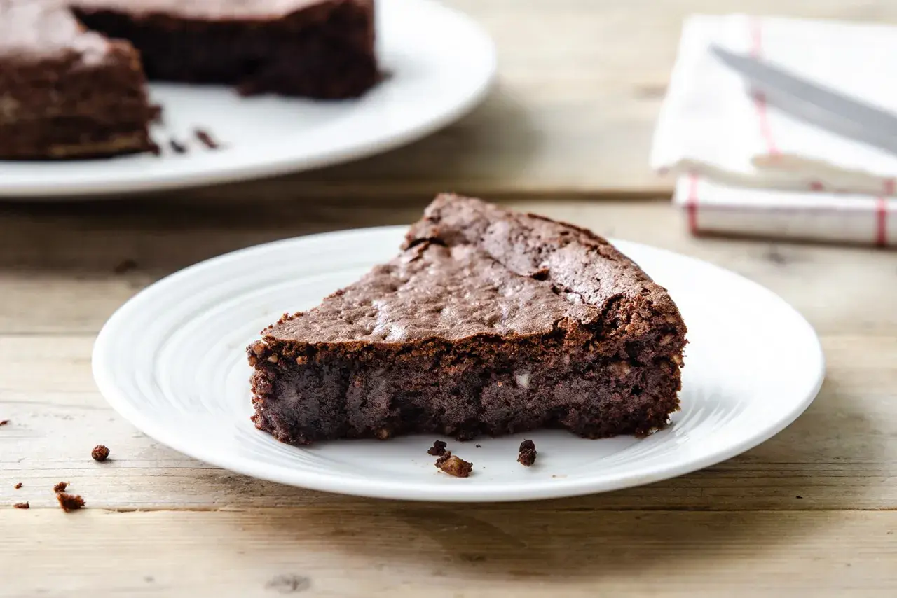 Una fetta di torta al cioccolato senza farina e burro, densa e umida, su un piatto bianco.