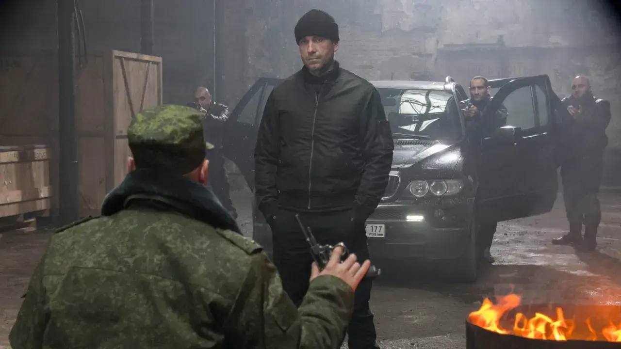 W magazynie, wśród dymu i ognia, mężczyzna w czapce i kurtce bomberce, niczym pitbull, ostatni pies, mierzy z pistoletu do żołnierza.