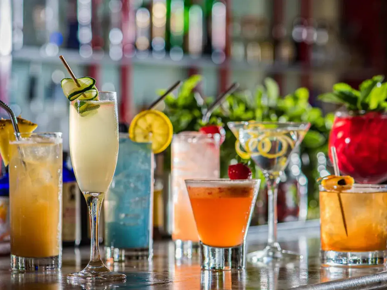 En rad färgglada drinkar, inklusive en sockerlag drink med gurka, står uppradade på en bardisk.