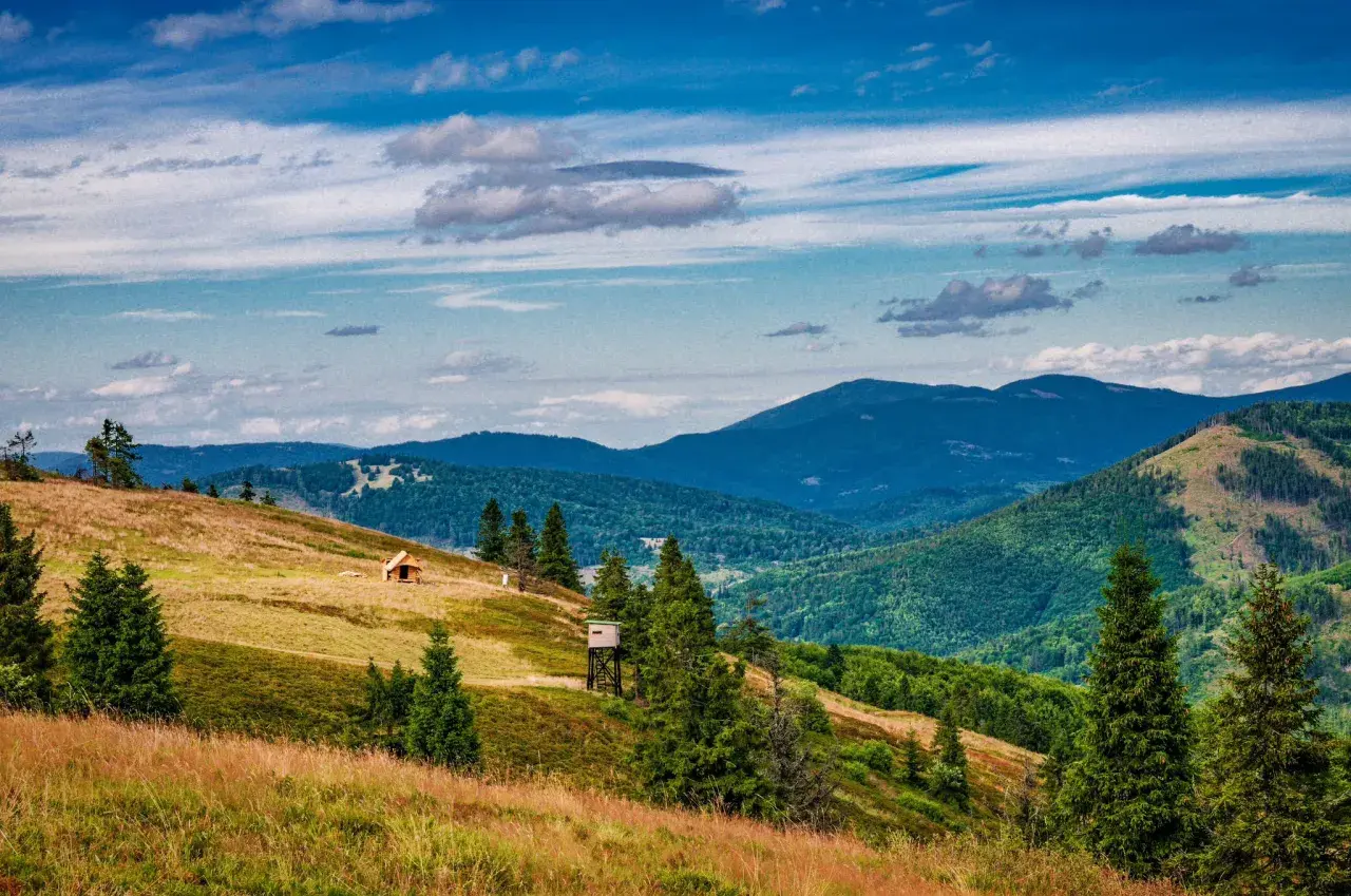 Widok na malowniczą **przełęcz Przegibek** z drewnianą chatką i wieżą widokową na zboczu. W tle gęsto zalesione góry pod błękitnym, chmurnym niebem.
