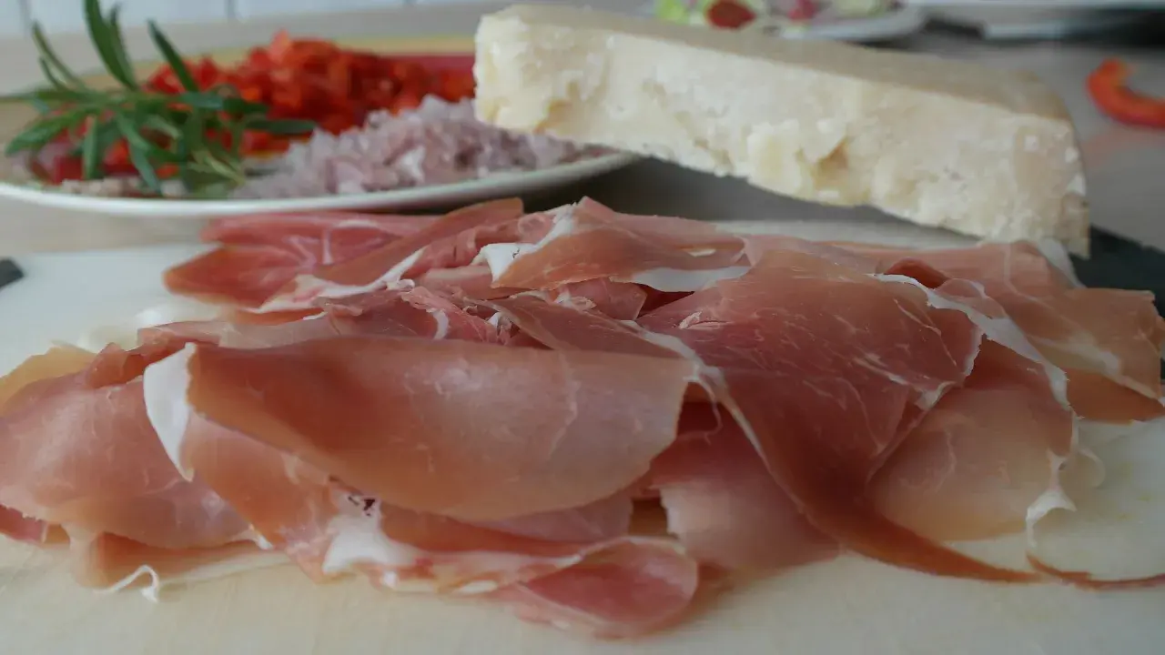 Cienko pokrojona szynka crudo, kawałek parmezanu i talerz z dodatkami.