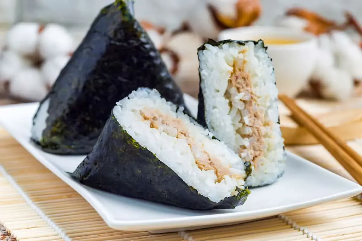 Three triangular onigiri triangle sushi with tuna filling, wrapped in nori seaweed, sit on a white plate with chopsticks and a cup of tea in the background.
