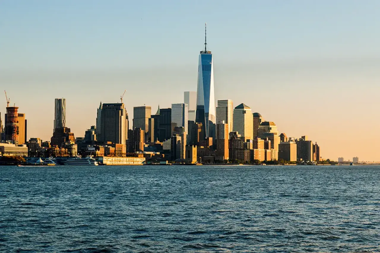 Widok na Manhattan z rzeką w pierwszym planie. W oddali widać drapacze chmur, w tym nowoczesne wieże World Trade Center, oświetlone zachodzącym słońcem.