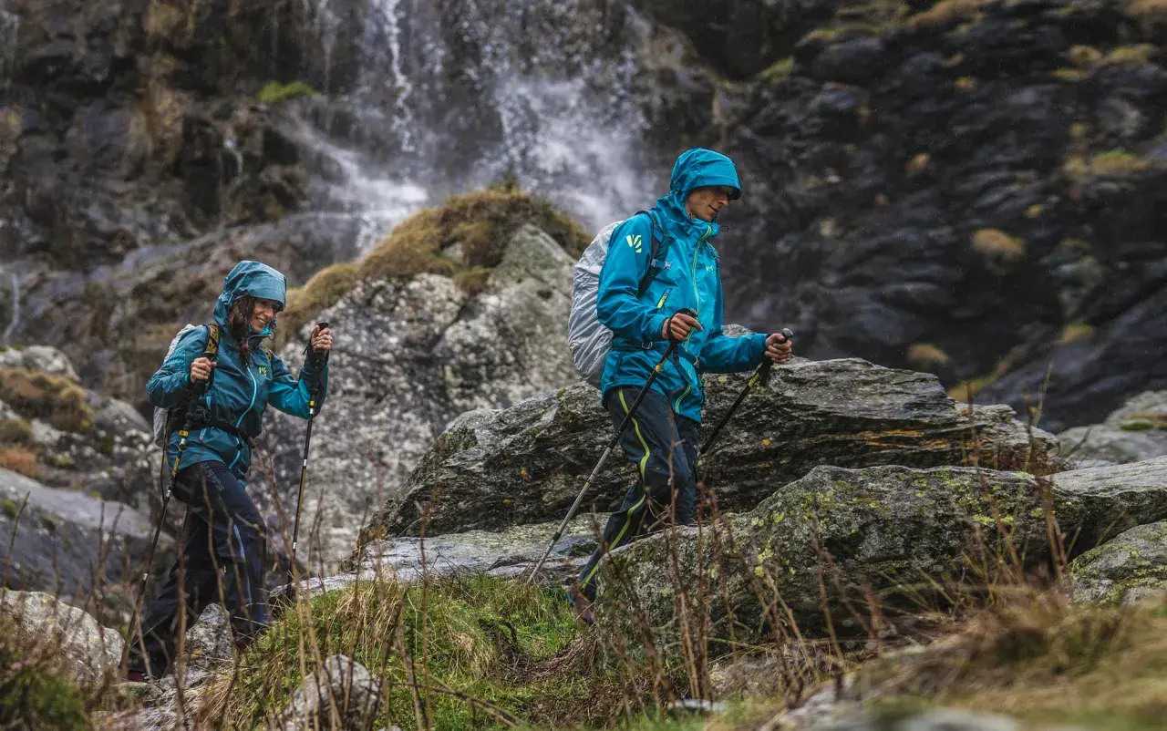 Dwie osoby w turkusowych kurtkach w góry, z kijkami trekkingowymi, idą obok wodospadu.