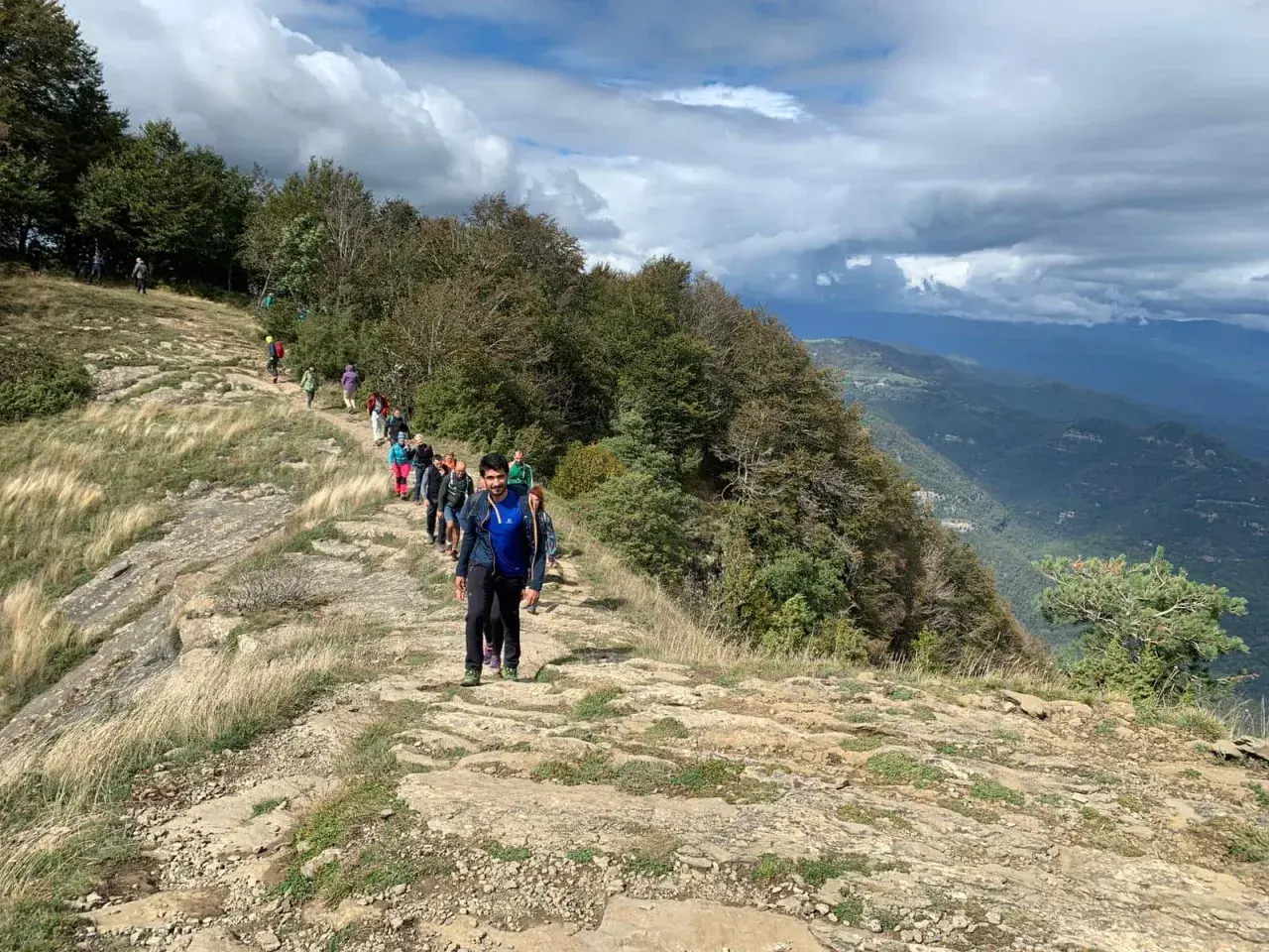 Grupo de excursionistas recorre la ruta puigsacalm, un sendero rocoso con vistas a un valle boscoso bajo un cielo nublado.