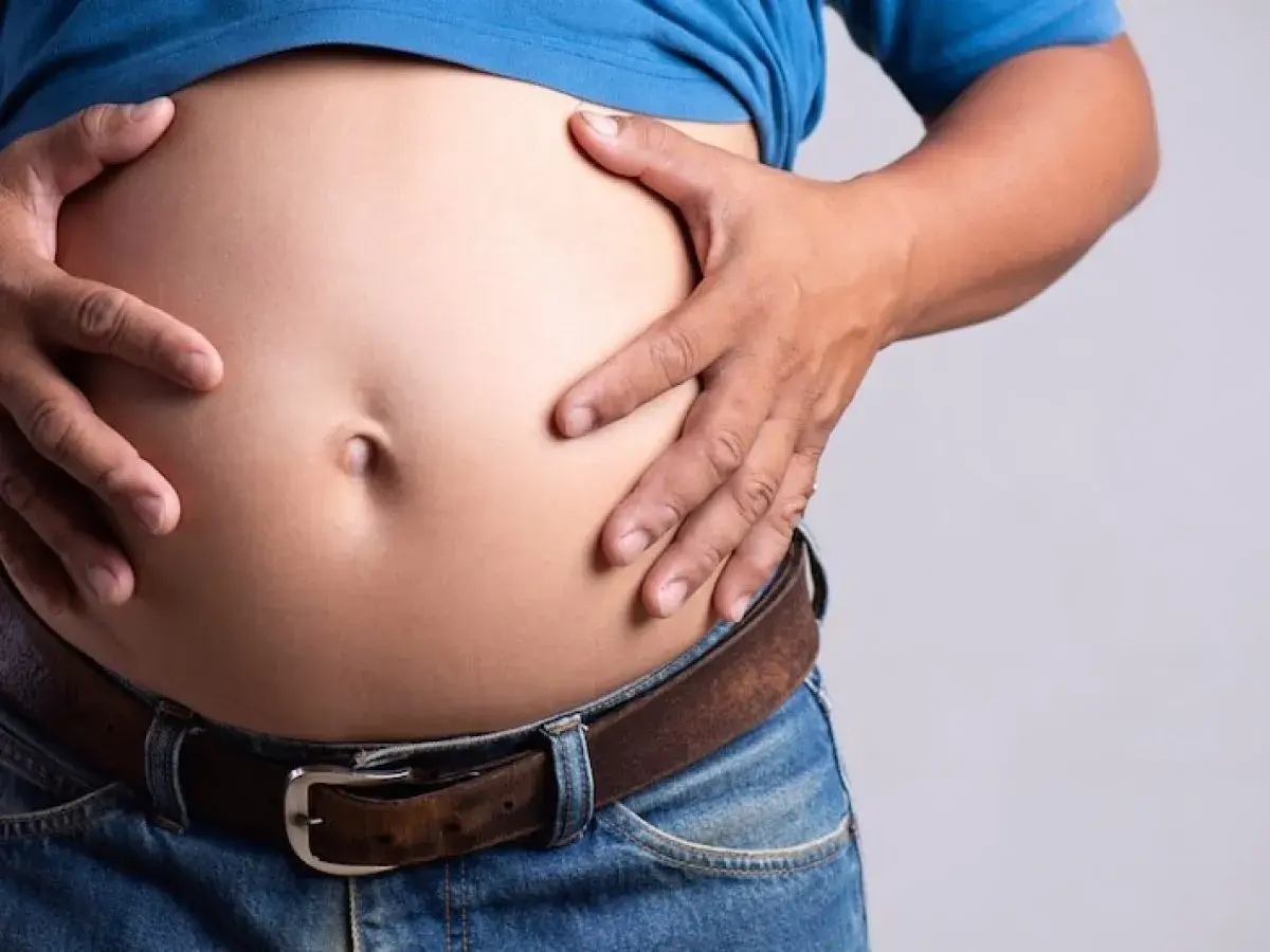 Un homme, je suis mince avec un gros ventre, tient son abdomen proéminent au-dessus de son jean et de sa ceinture.