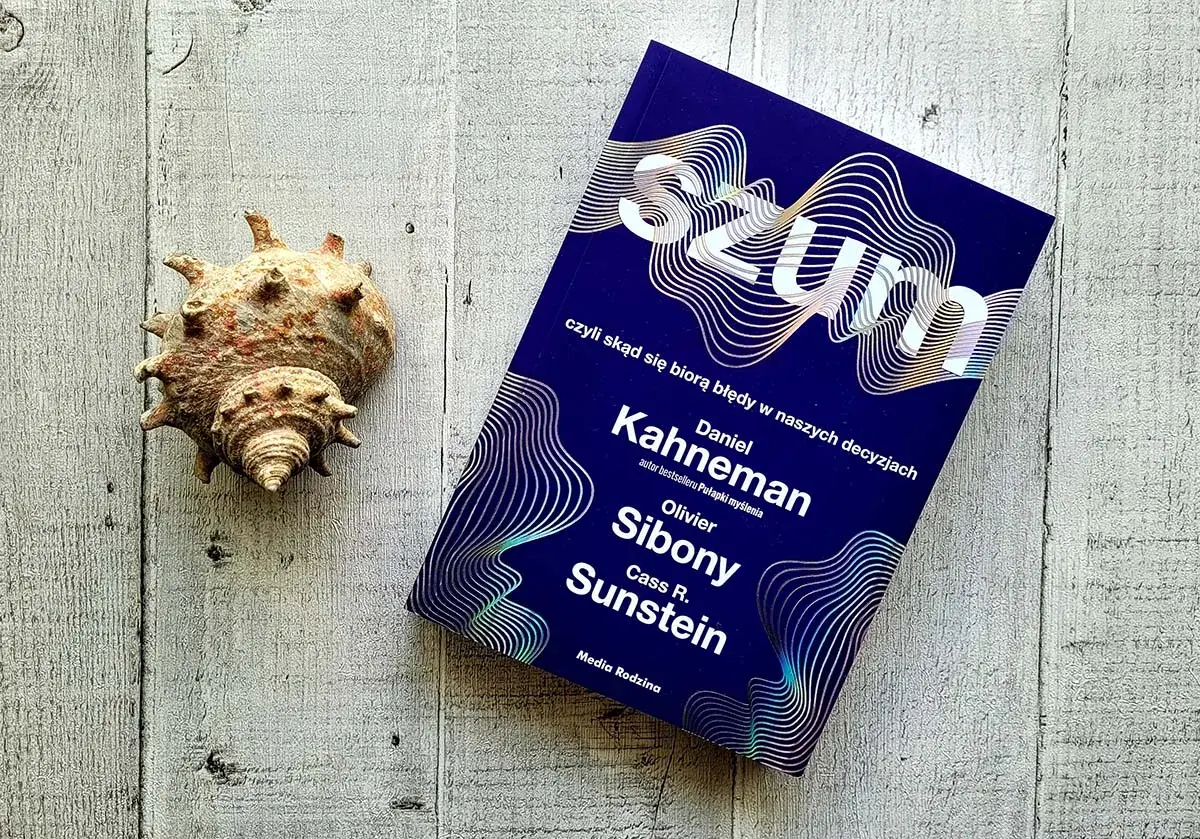 Książki Daniela Kahnemana, "Szum", obok muszli na drewnianym tle.
