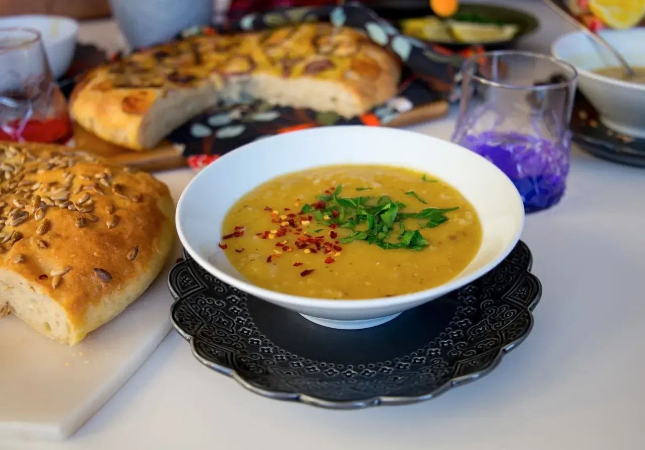 En skål med varm shorbet adas, toppad med persilja och chiliflakes, bredvid ett solrosbröd och focaccia.
