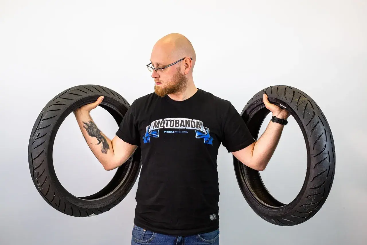 Mężczyzna w koszulce "Motobanda" trzyma dwa motocyklowe opony, zastanawiając się, ile lat mogą mieć opony.