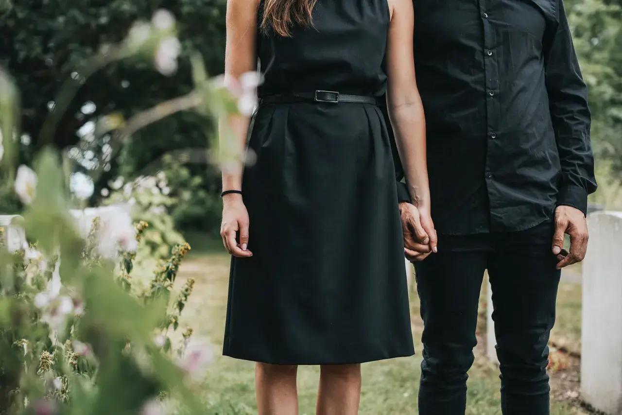 Ein Paar in schwarzer Kleidung, die Frau in einem schlichten Kleid, hält Händchen. Passende **Beerdigung Kleidung Frau Sommer** für einen traurigen Anlass.