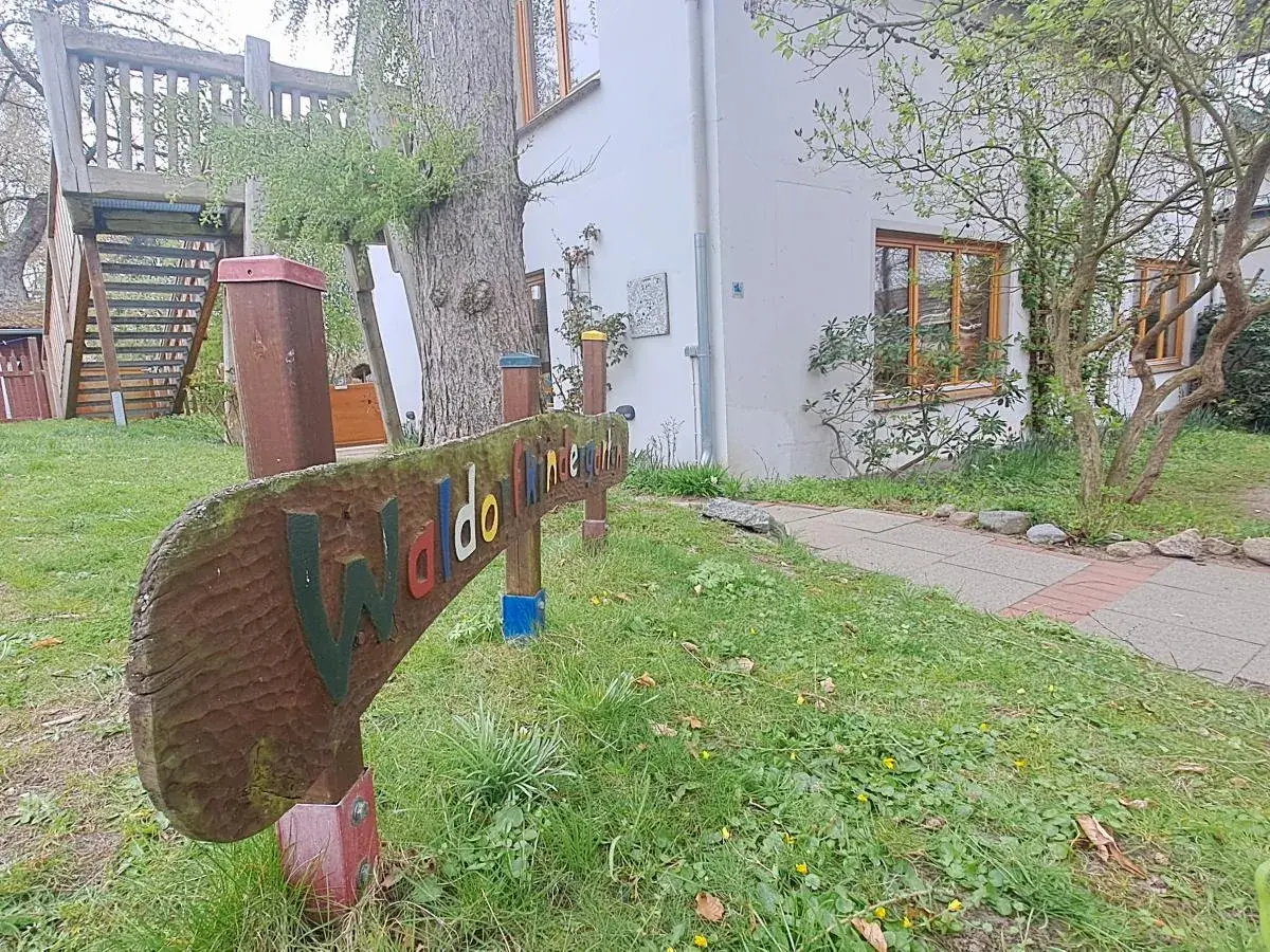 Holzschild "Waldorfkindergarten" vor einem Gebäude mit Spielturm. Mögliche Nachteile: wenig Struktur, viel Freiheit.