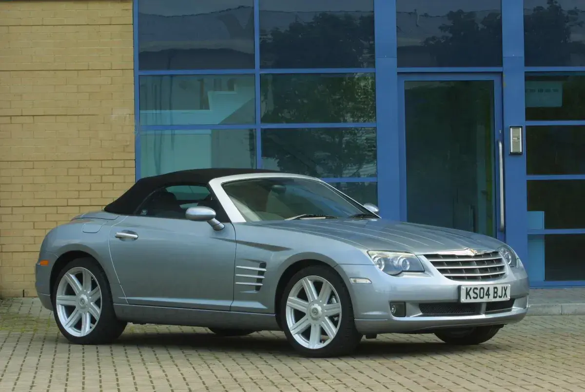 Srebrny Chrysler Crossfire auto z czarnym dachem stoi przed budynkiem z żółtej cegły i niebieskimi oknami.
