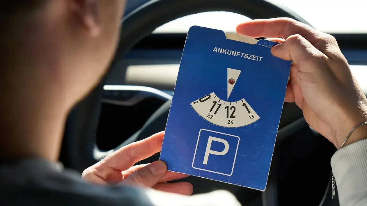 Person hält eine Parkscheibe mit der Aufschrift "Ankunftszeit" und dem Parkplatzsymbol.