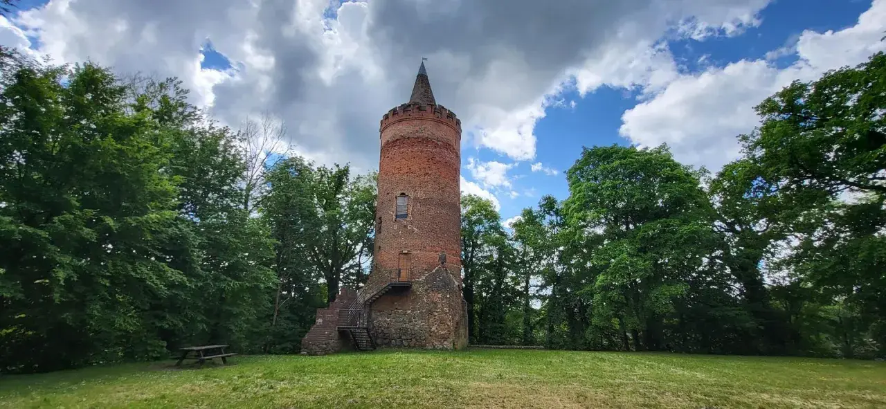 Stara, ceglana wieża zamkowa z kamiennym fundamentem, otoczona zielenią drzew, pod błękitnym niebem z chmurami.