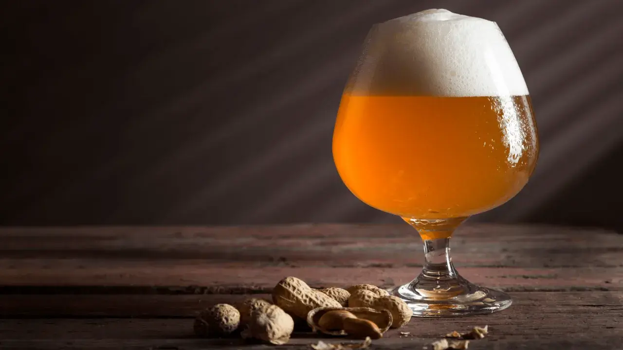 Un bicchiere di keller beer dorata con schiuma bianca, accompagnata da arachidi su un tavolo di legno scuro.