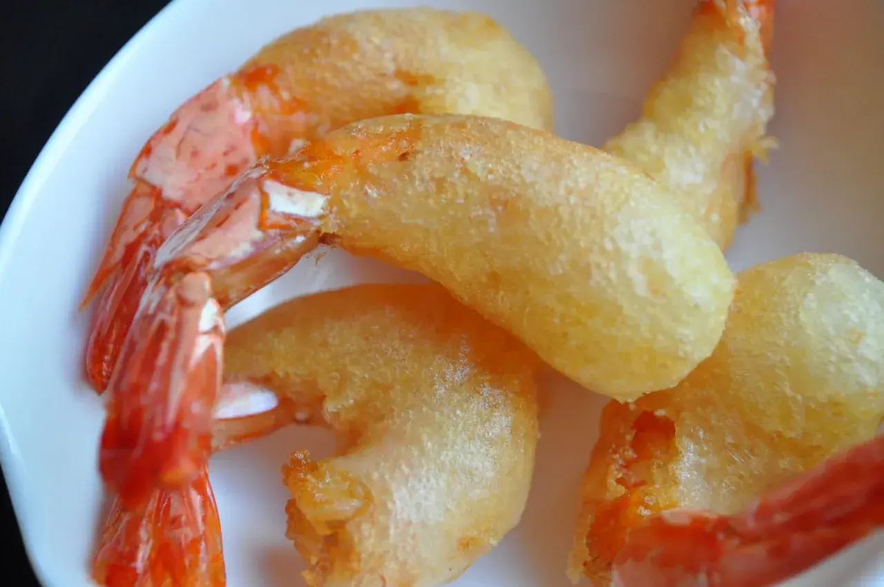 Plusieurs beignets de crevette dorés et croustillants, avec des queues de crevettes roses qui dépassent, servis dans une assiette blanche.