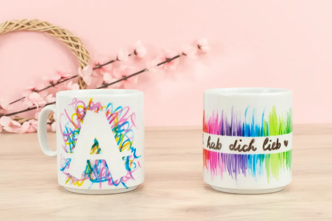Kreative tassen bemalen ideen: eine Tasse mit dem Buchstaben A und bunte Kritzeleien, daneben eine Tasse mit Regenbogenfarben und der Aufschrift "hab dich lieb".