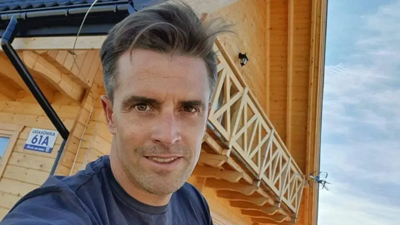 Staszek Karpiel-Bułecka pozuje na tle drewnianego domu z balustradą.