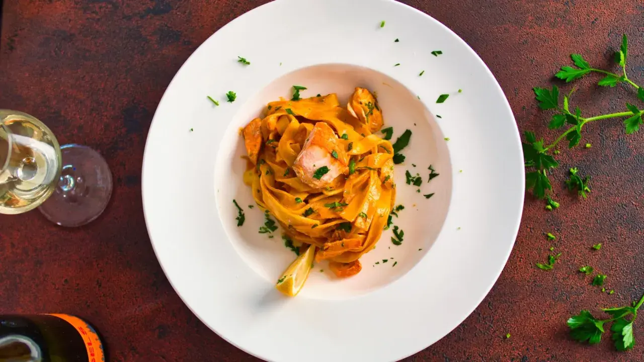 Ein Teller Tagliatelle al Salmone, garniert mit Petersilie und einer Zitronenspalte, daneben ein Glas Weißwein.