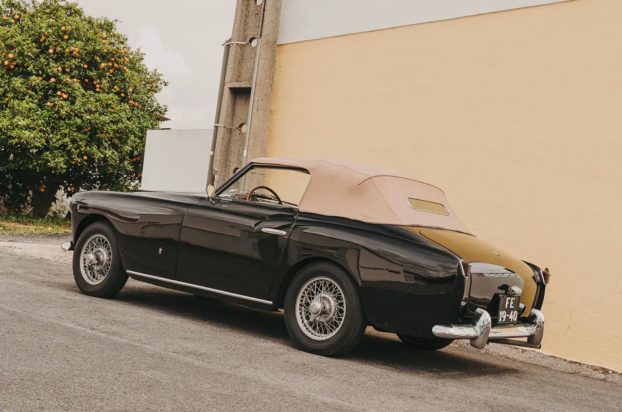 Svart, klassisk brittisk sportbil med beige sufflett parkerad vid en gul vägg, bredvid ett apelsinträd.