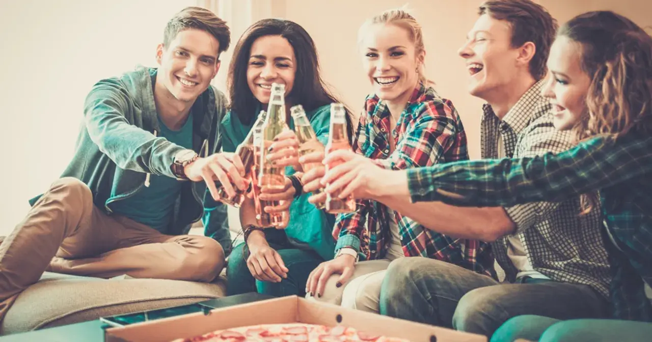 Grupa przyjaciół wznosi toast butelkami piwa, śmiejąc się i jedząc pizzę. Nawet trudne pytania nie psują im nastroju.