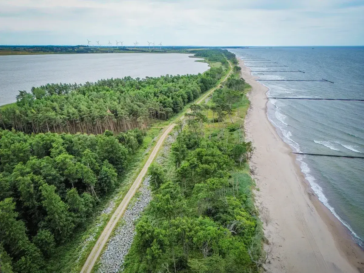 Malownicza nadmorska trasa rowerowa biegnie wzdłuż lasu i plaży, z widokiem na morze i jezioro.