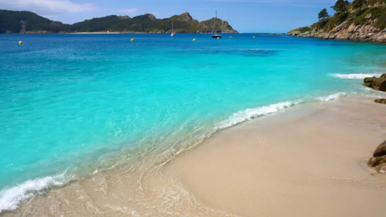 Aguas turquesas y arena dorada en unas playas cristalinas España, con veleros navegando al fondo y montañas verdes.