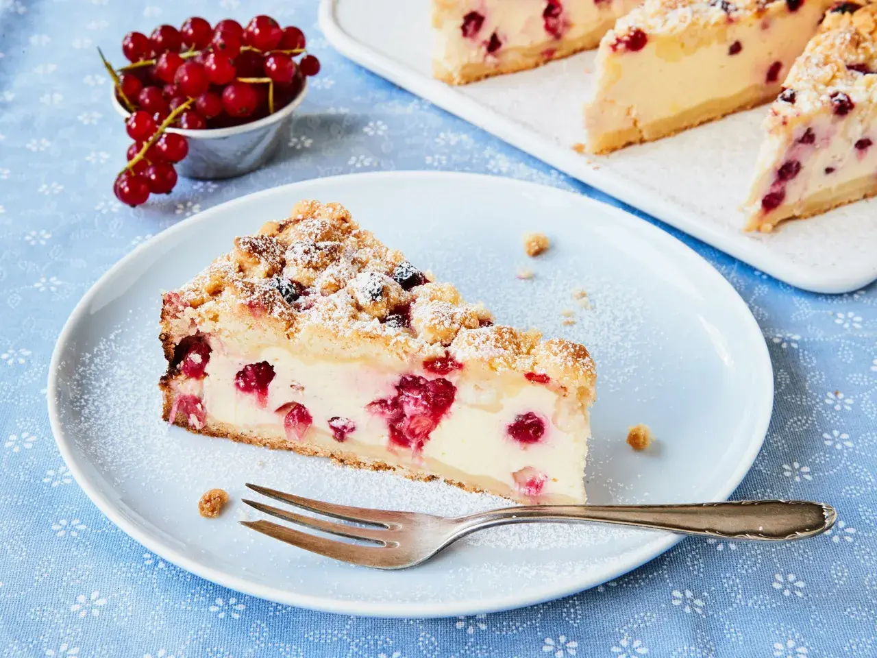 Ein Stück Johannisbeerkuchen mit Quark, bestäubt mit Puderzucker. Frische rote Johannisbeeren liegen daneben.