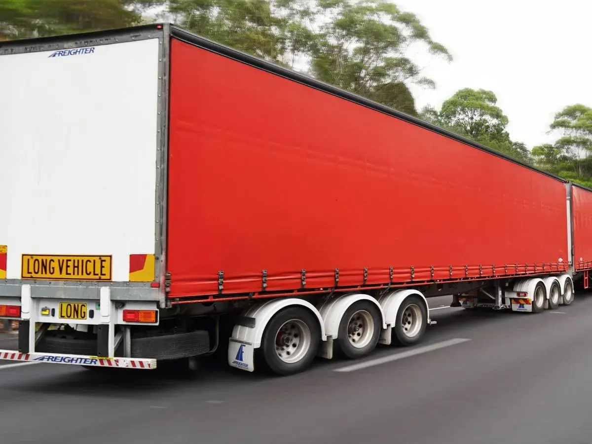 Un long camion 60 tonnes, avec une remorque rouge vif, roule sur une route bordée d'arbres.