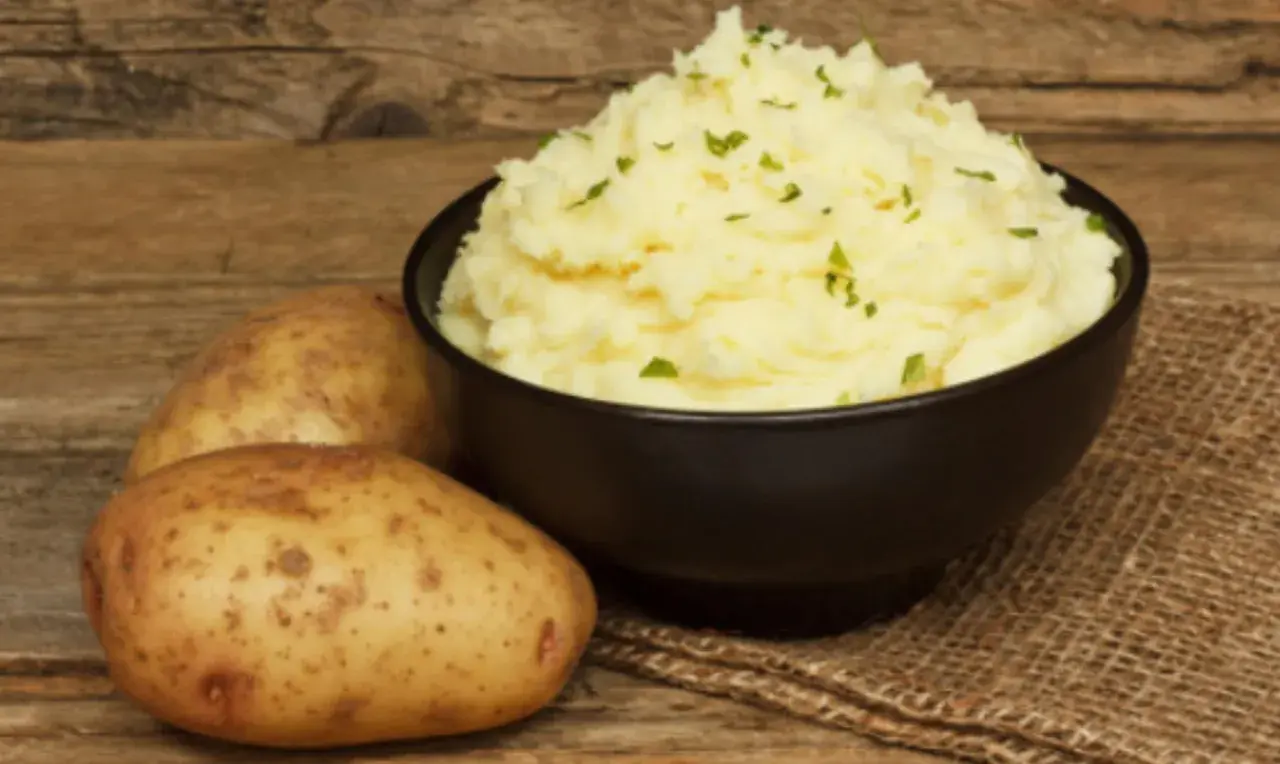 Un cremoso puré de patatas Thermomix, adornado con perejil fresco, junto a patatas crudas sobre una mesa de madera.