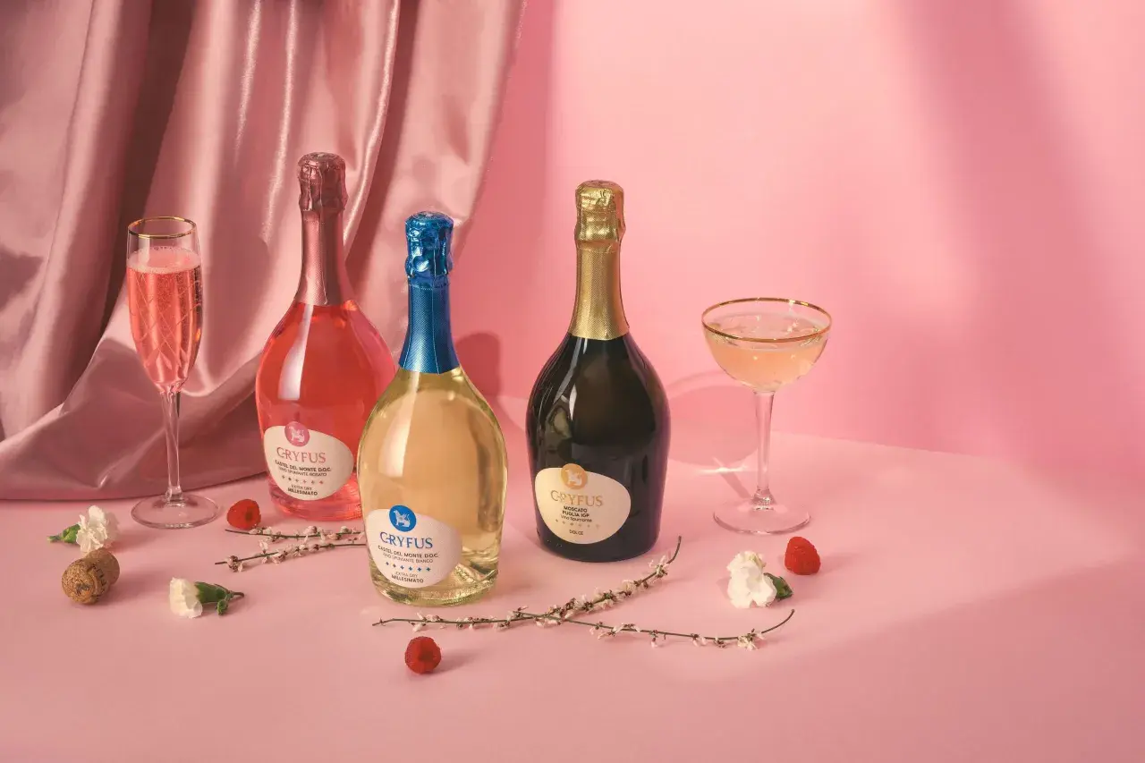 Tre bottiglie di champagne italiano, una rosata, una bianca e una dorata, con calici e decorazioni floreali su sfondo rosa.