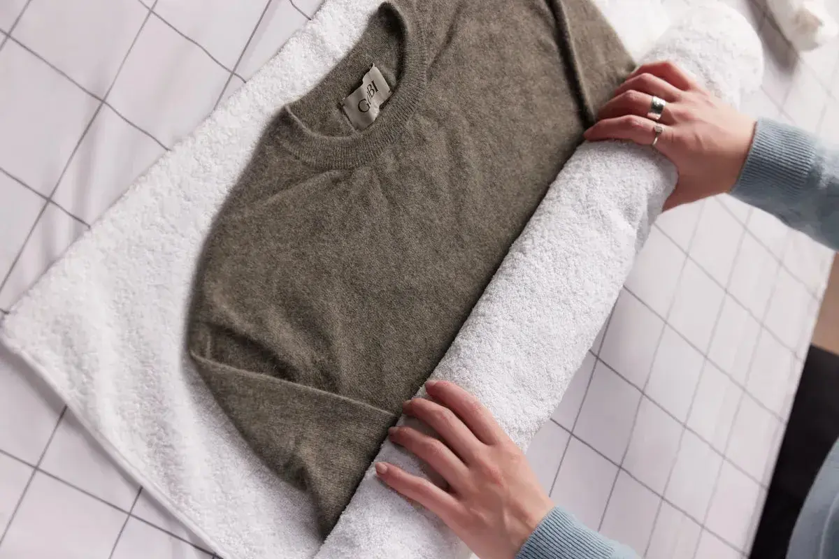 Hände rollen einen braunen Kaschmirpullover auf einem weißen Handtuch auf, um ihn nach dem Kaschmir waschen zu trocknen.