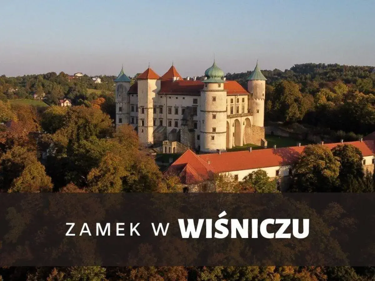Majestatyczny zamek Wiśnicz otoczony jesiennymi drzewami, z charakterystycznymi wieżami i czerwonymi dachami.