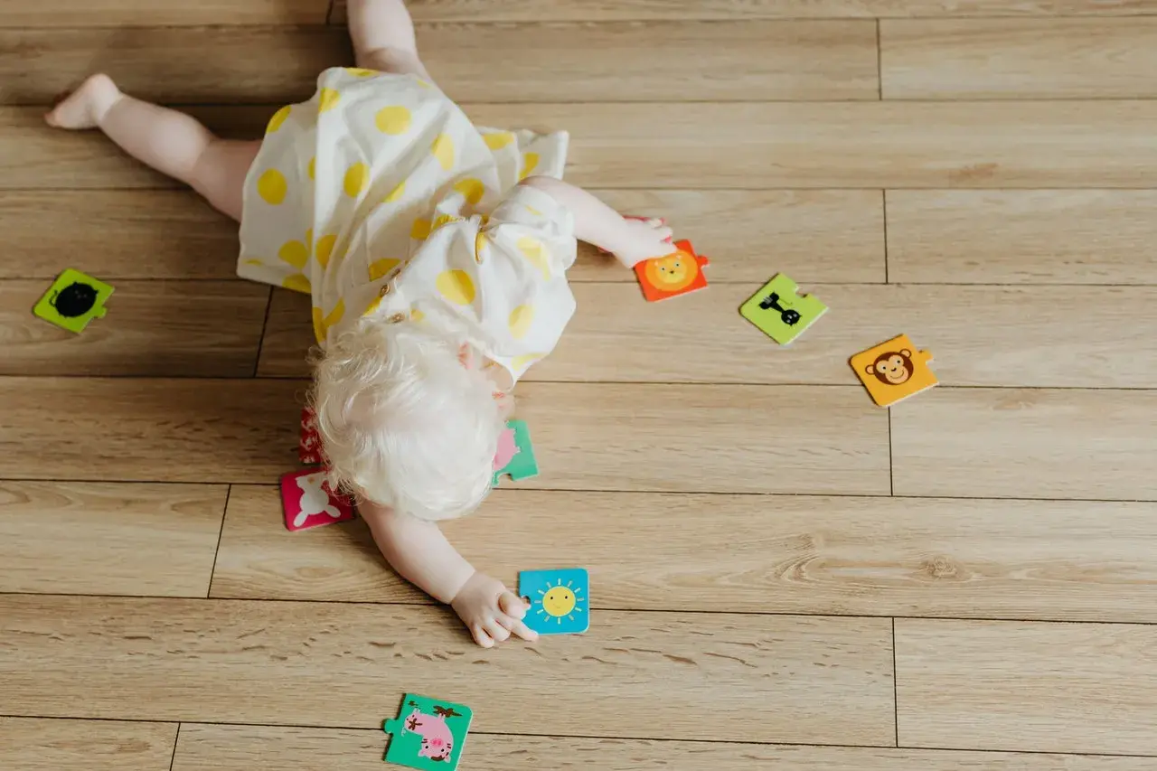 Dziecko z jasnymi włosami, w żółtej sukience w kropki, przewraca się na brzuch wśród kolorowych puzzli z zabawnymi zwierzątkami i słońcem.