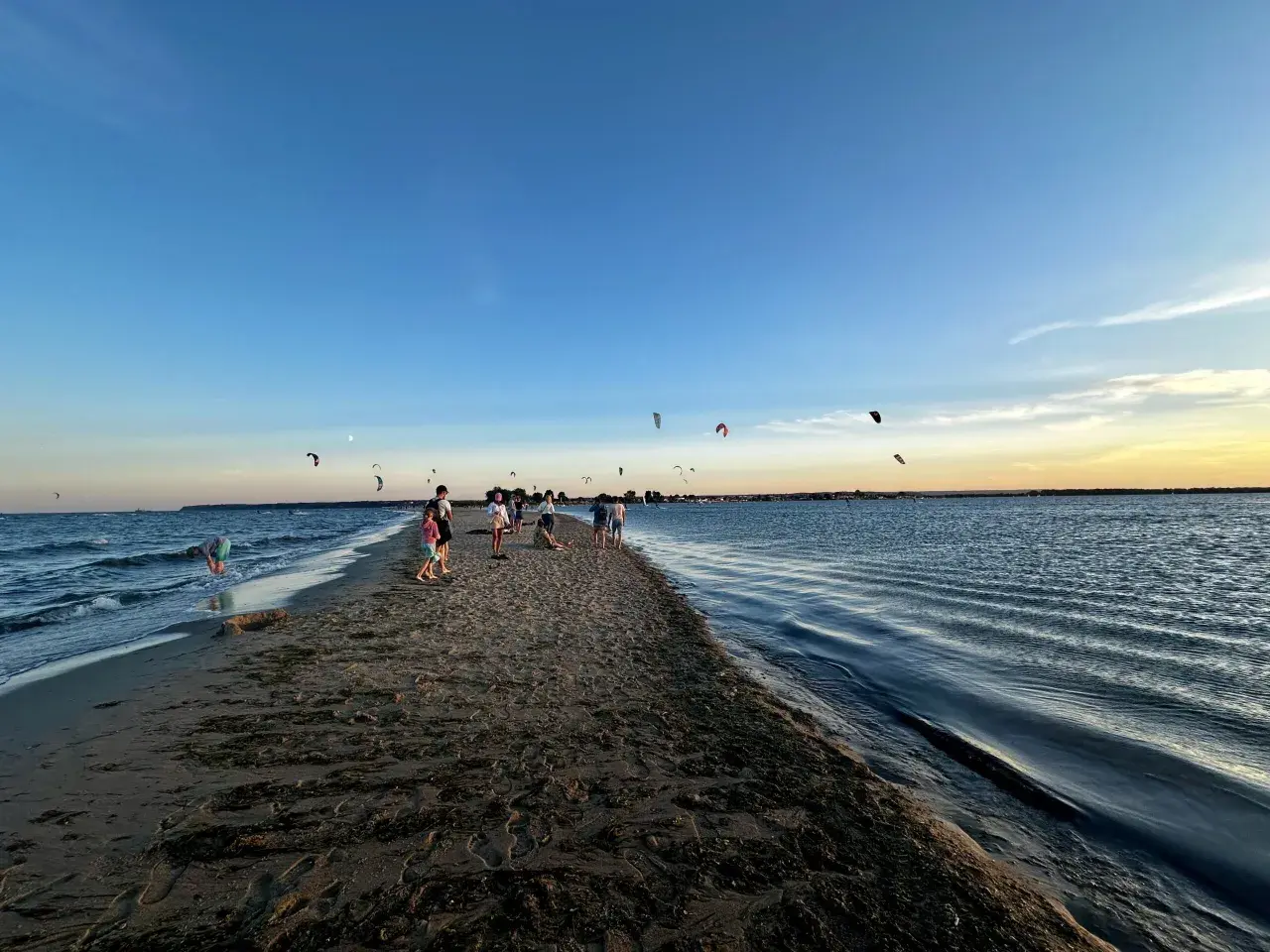 Ludzie spacerują po piaszczystym cyplu Rewskim, gdzie można podziwiać kitesurferów. Jak dojechać? To proste, wystarczy kierować się nad morze.