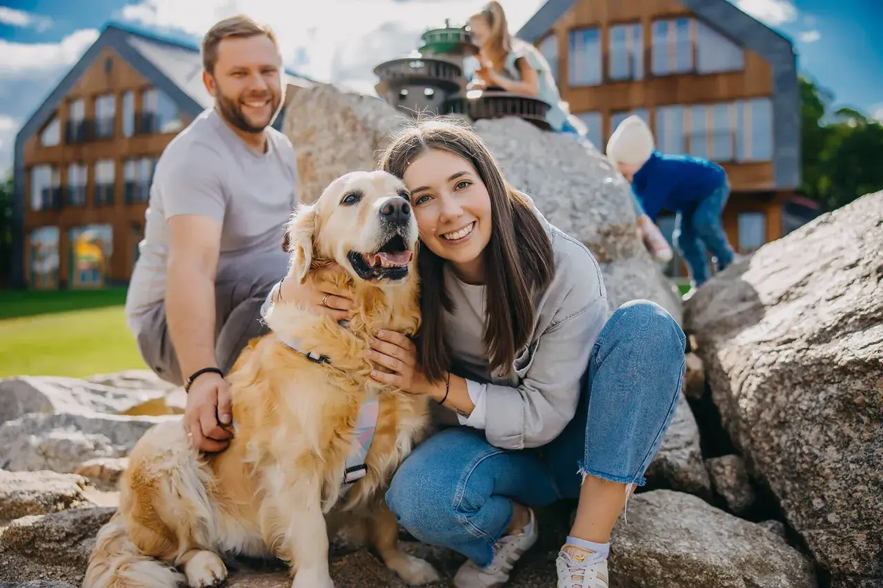 Uśmiechnięta rodzina z psem na tle drewnianych domków. Czy na Śnieżkę można z psem? Tak, jeśli jest tak szczęśliwy jak ten złoty retriever!